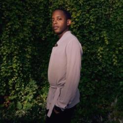 A person with short hair stands outdoors against a lush green wall of foliage. They are wearing a light-colored, long-sleeve shirt and are looking slightly to the side with a calm expression. The lighting casts a soft shadow, creating a warm, natural ambiance.